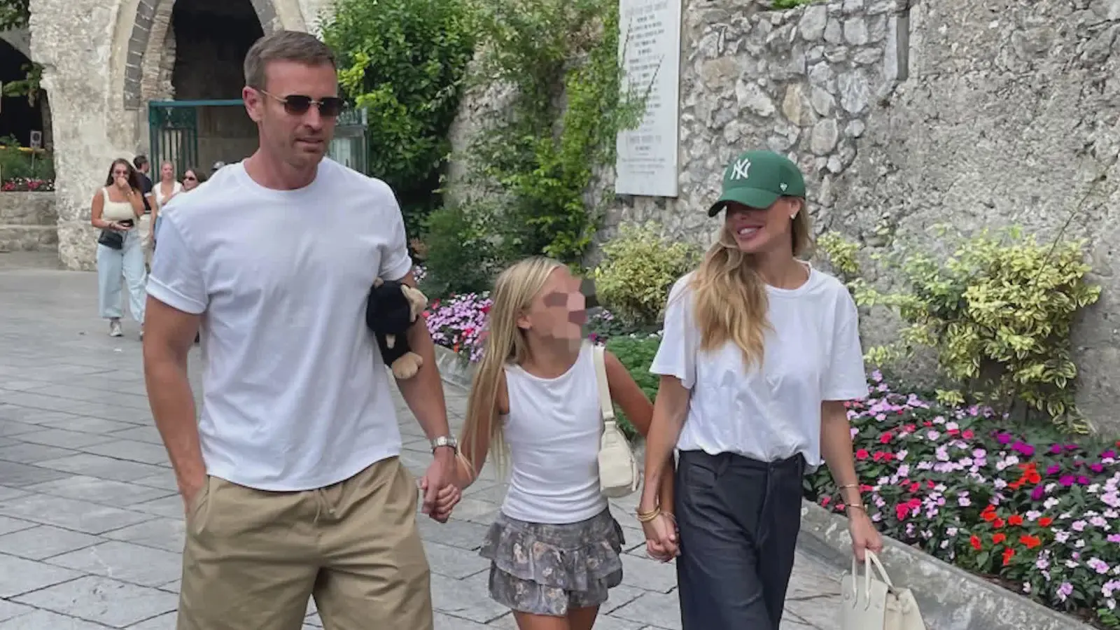 Ilary Blasi e Bastian Müller tengono per mano una bambina con il volto oscurato, lungo un percorso fiorito.